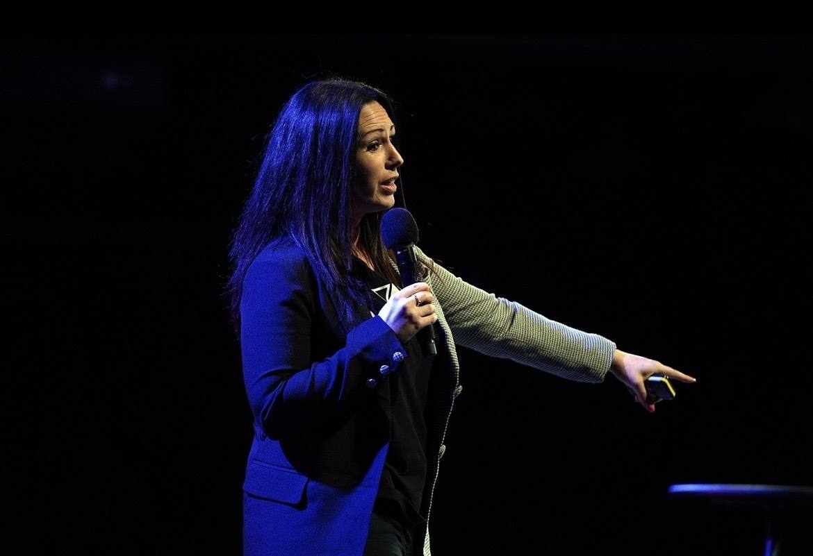 Sadie Restorick speaking on a dark stage, pointing into the audience